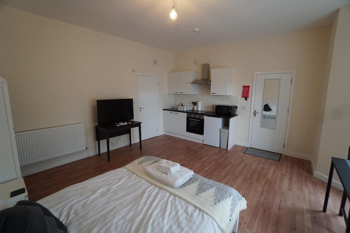 A hotel room with a bed, a TV, a kitchenette, and a door with a red sign.