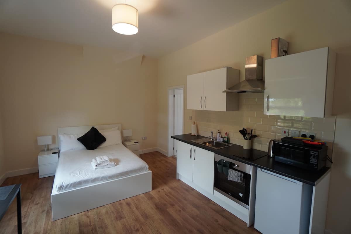 A compact hotel room with a bed, bedside table, and a kitchenette including a microwave and oven.