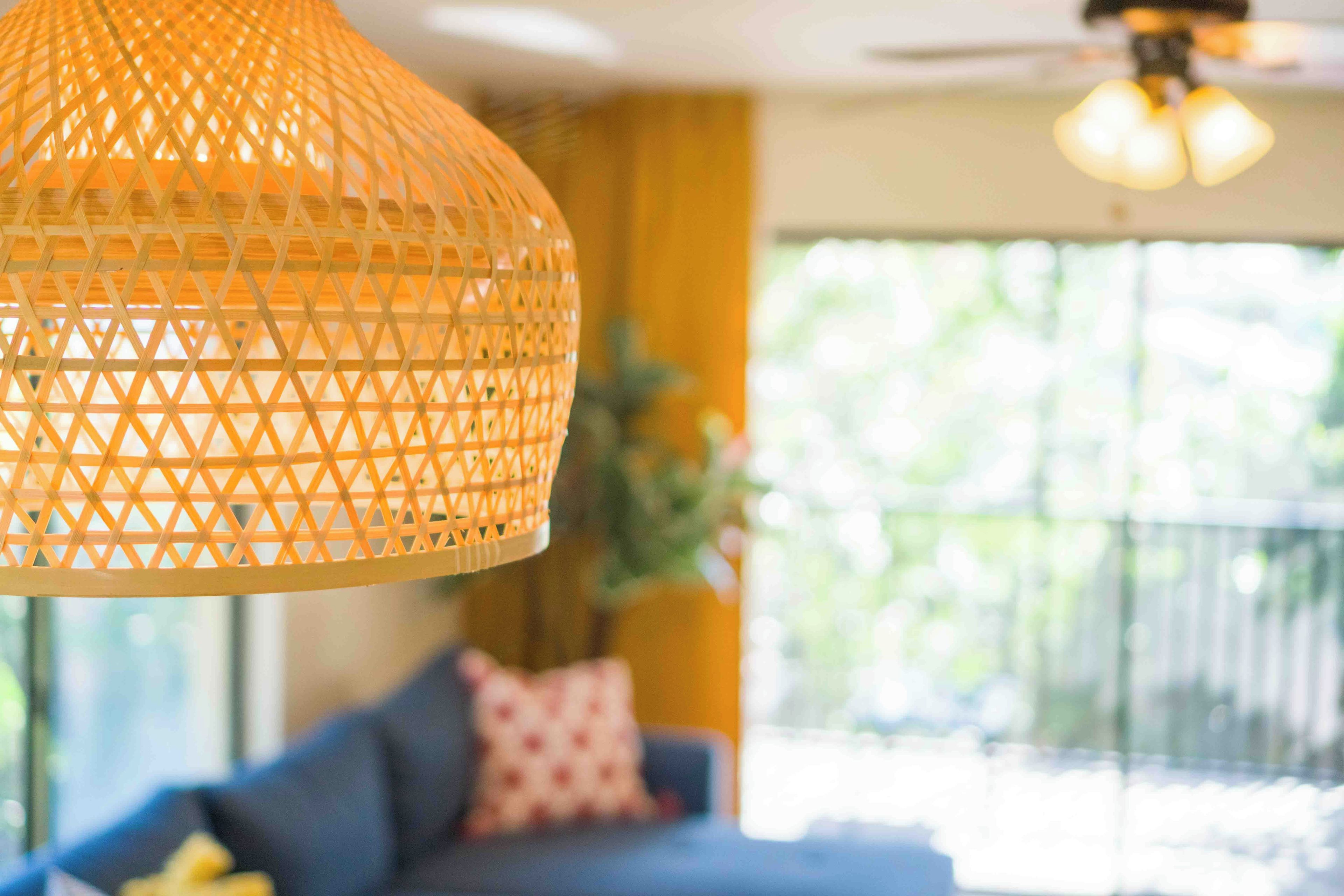 A room with a hanging woven light fixture, a ceiling fan, a sofa with a cushion, and a window with a view of greenery.