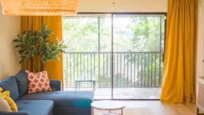 Sala de estar com sofá, uma mesinha, planta em vaso e vista para uma varanda com cortinas amarelas.