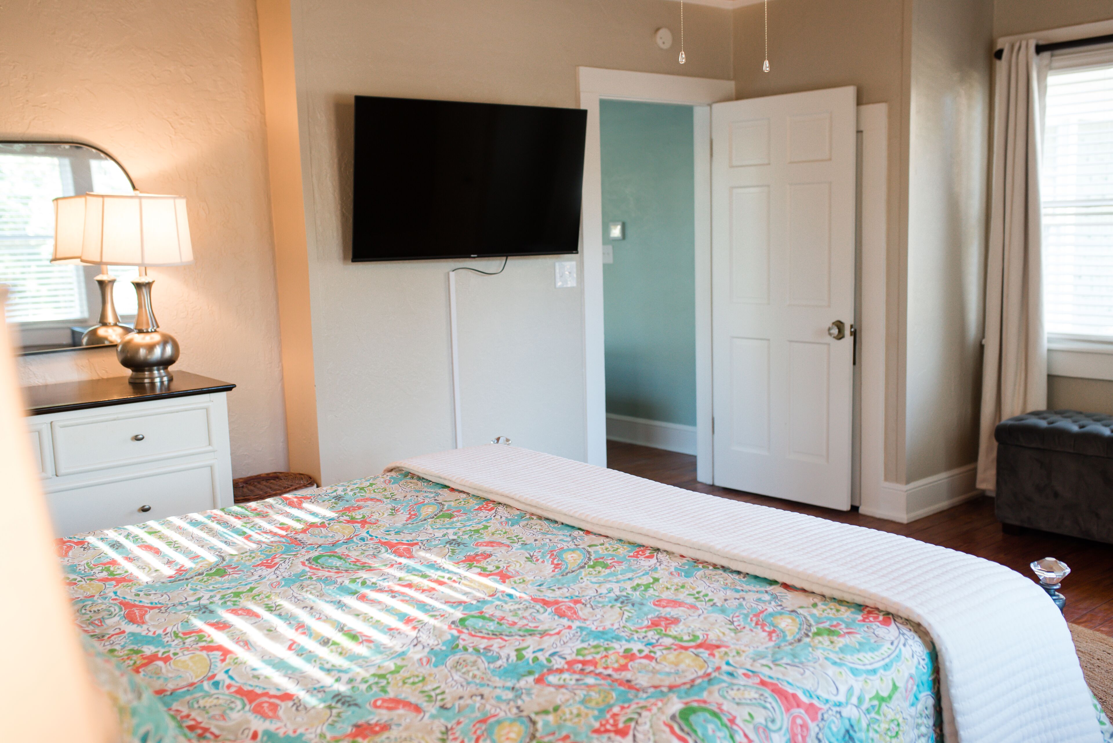 A bedroom with a bed, a dresser, a television mounted on the wall, and a door leading to another room.