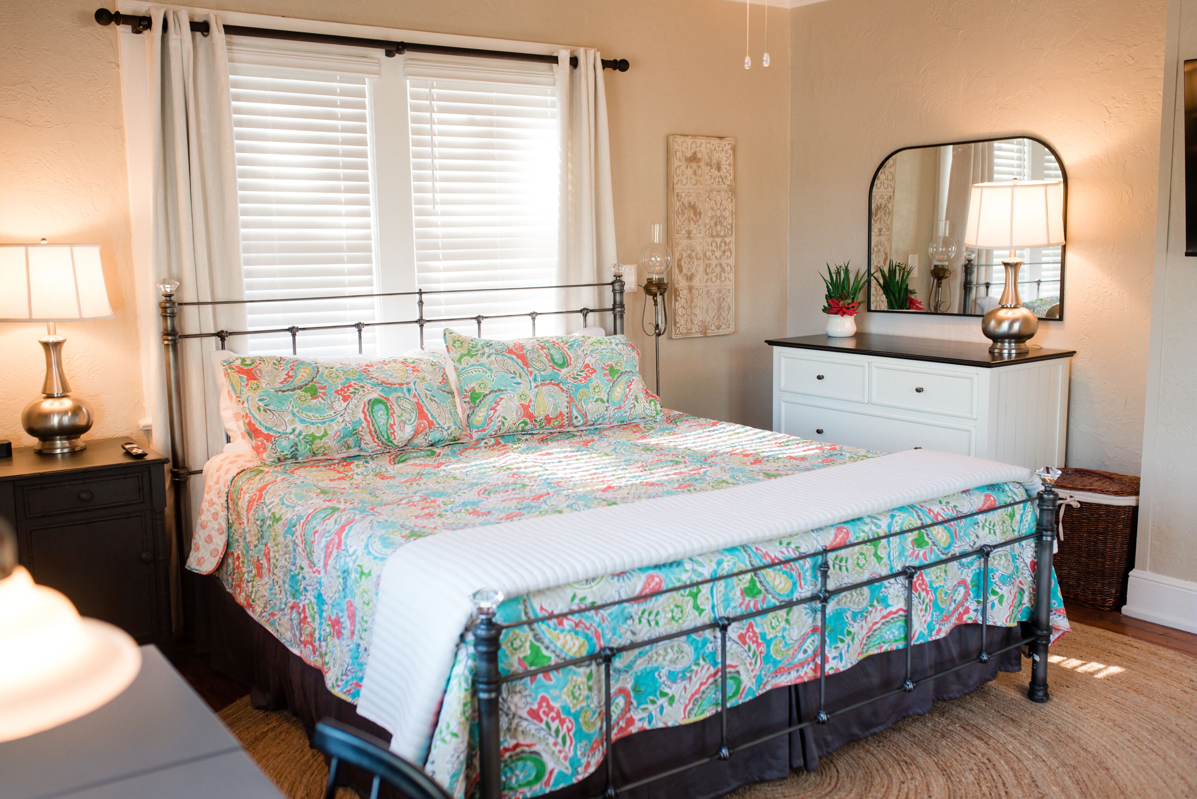 Un dormitorio con cama, mesitas de noche, cómoda, espejo y ventana con cortinas.