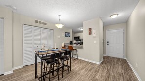 Una cocina moderna con armarios blancos, microondas y cafetera. Hay un comedor con una mesa de madera y taburetes de bar.