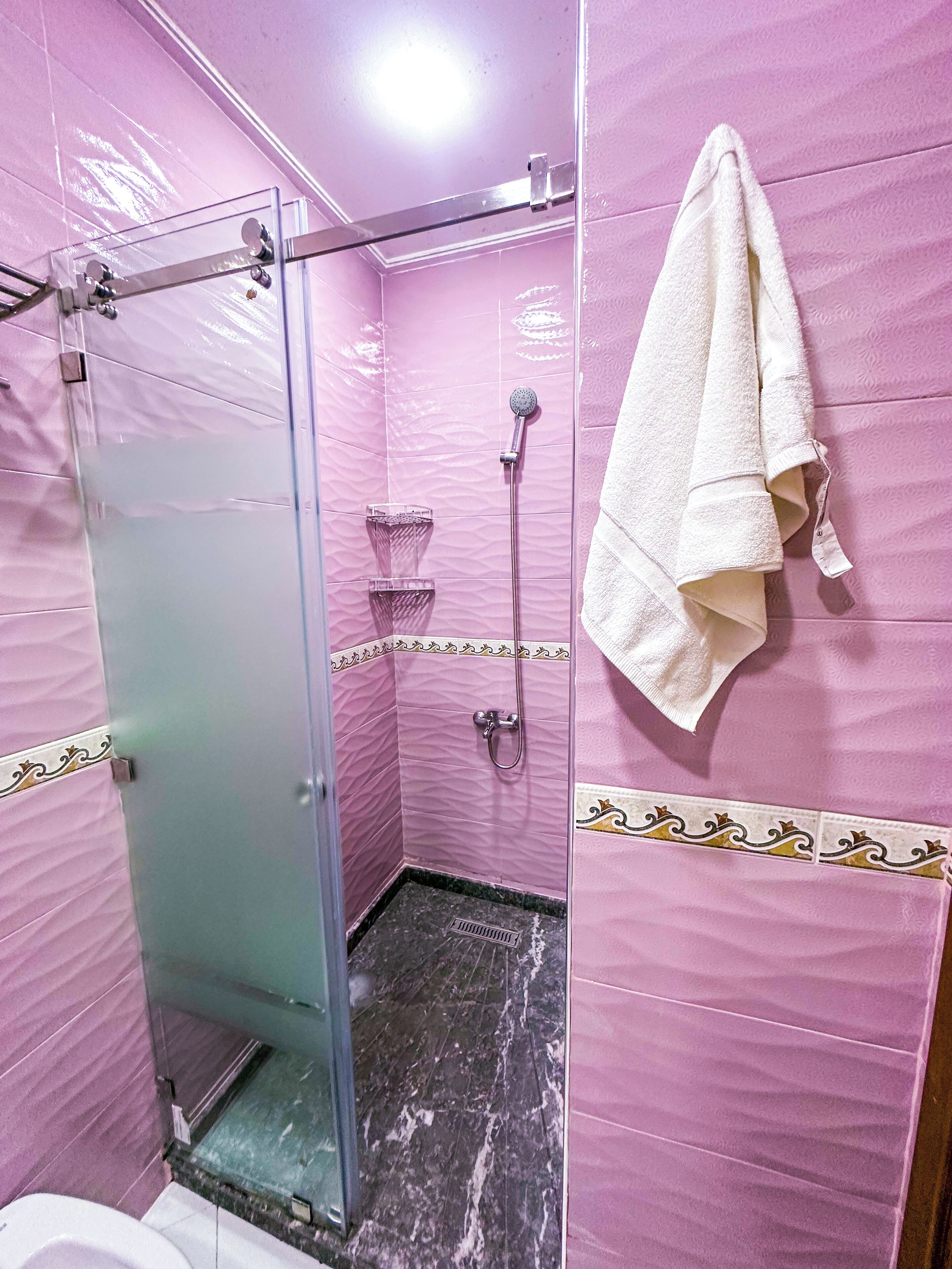 A bathroom with pink tiled walls, a glass shower enclosure, a white towel hanging on the wall, and a marble floor.