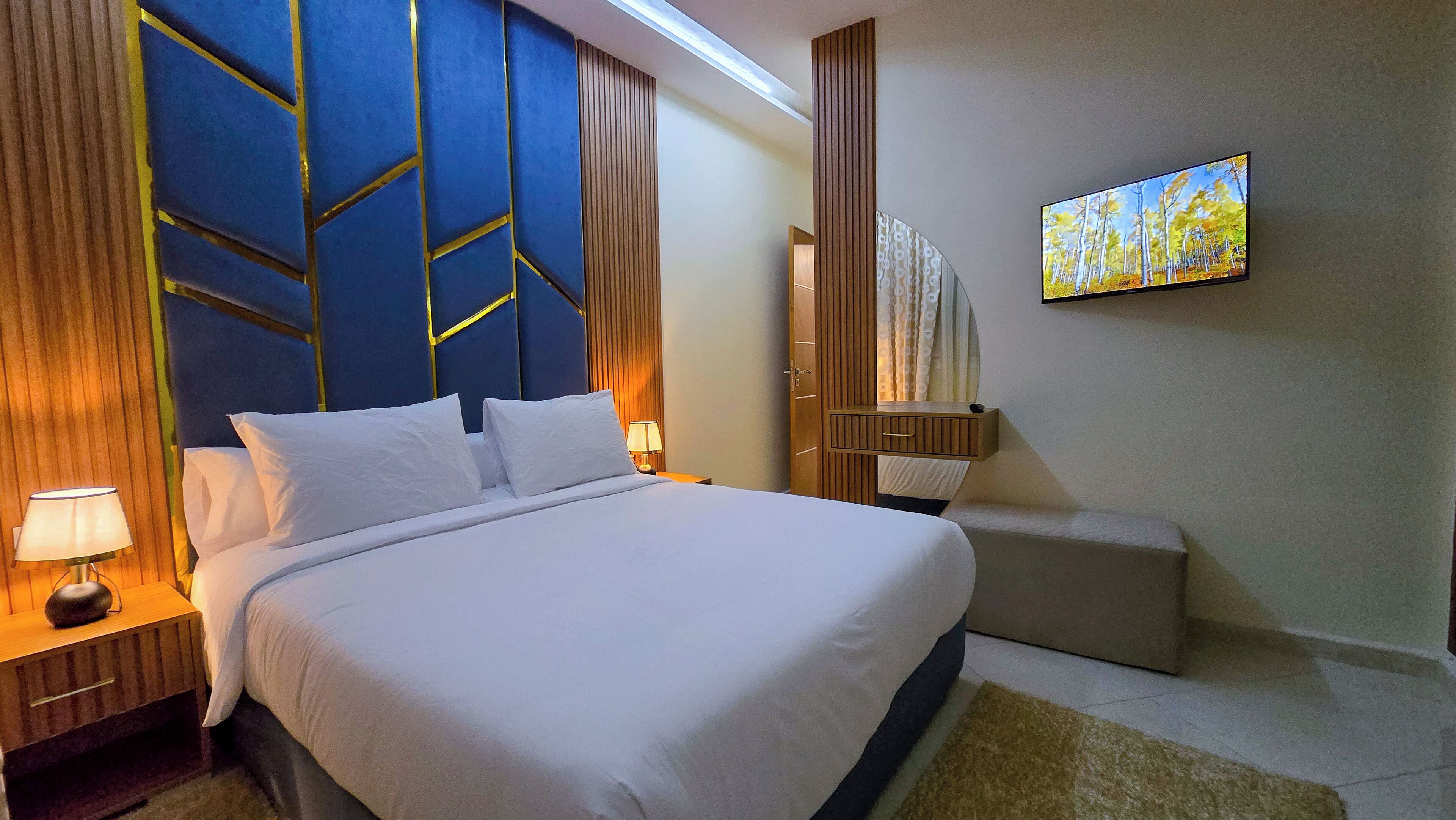 Ein Hotelzimmer mit einem großen Bett, einem Nachttisch mit Lampe, einem Holzbeistelltisch, einer kleinen Sitzecke und einem an der Wand befestigten Fernseher, der eine Waldszene zeigt.