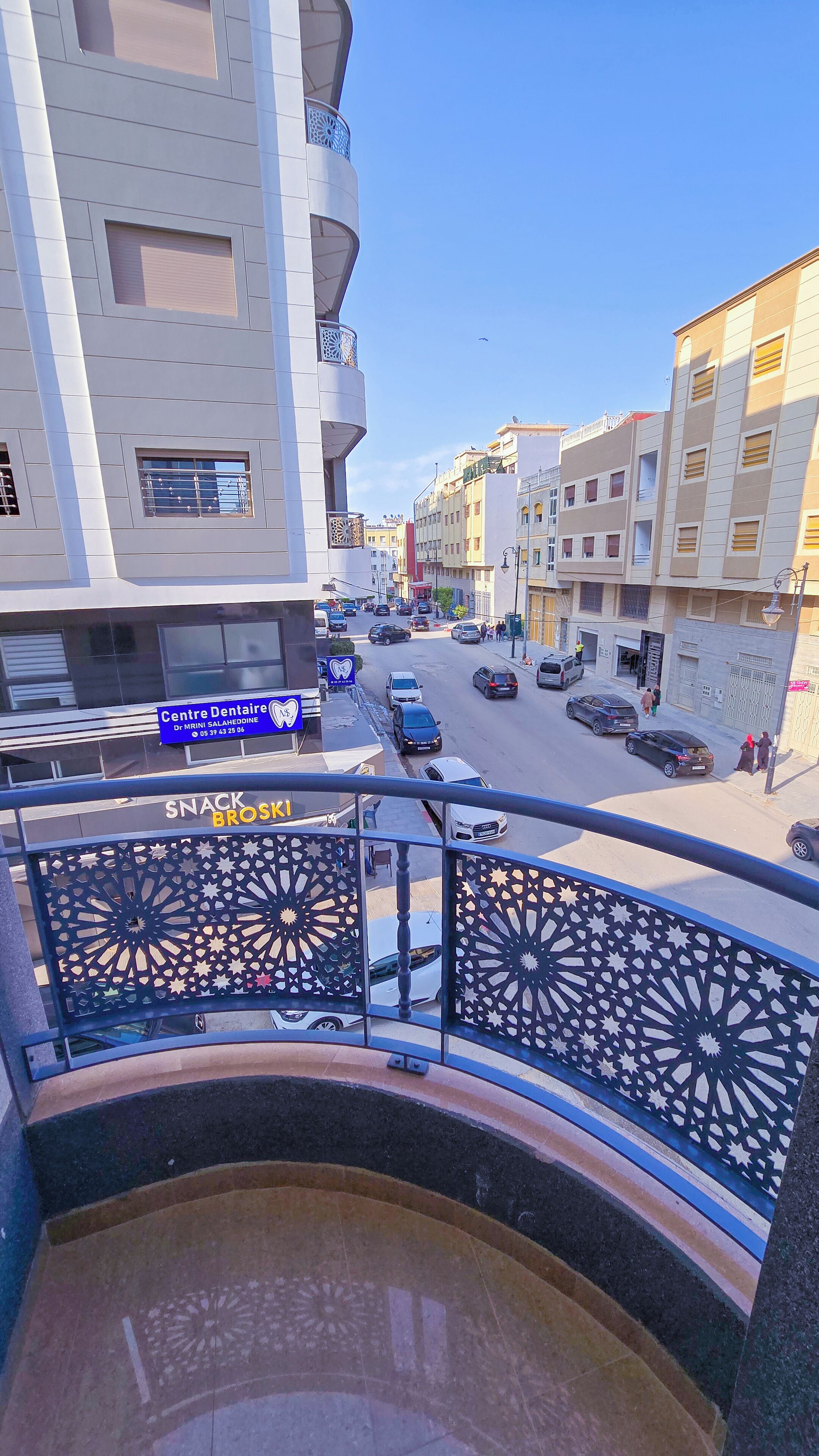 A street view from a balcony with a railing, buildings, cars, and a sign for a dental center.