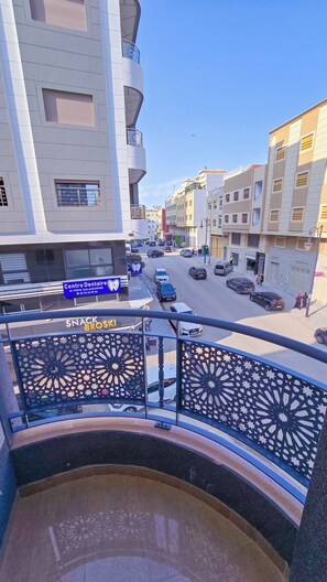 Vista de la calle desde un balcón con barandilla decorativa, edificios y coches estacionados.