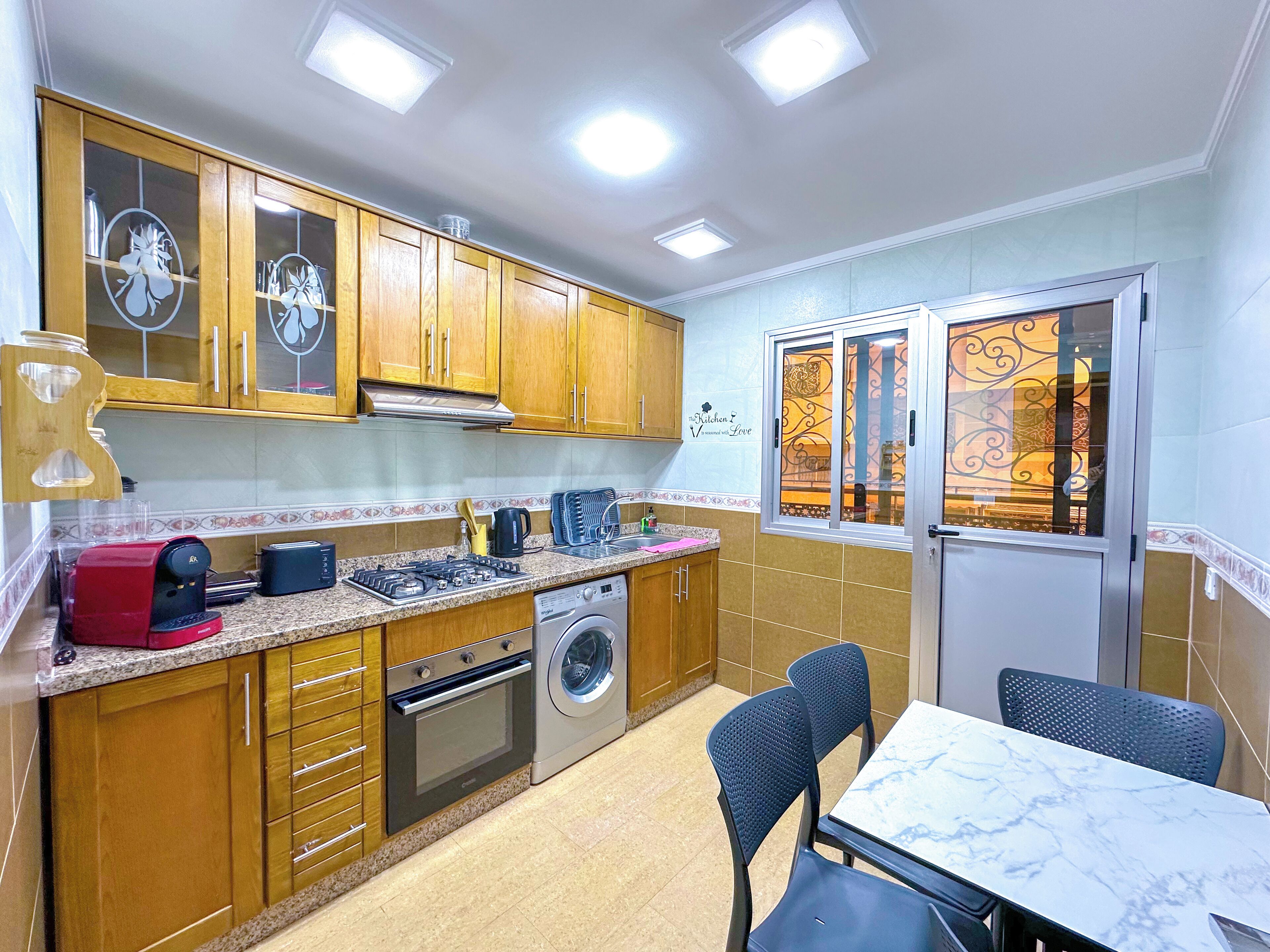 Cocina compacta con armarios de madera, estufa a gas, lavadora y comedor con mesa y sillas.