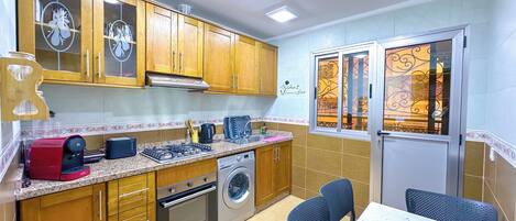 Cocina compacta con armarios de madera, estufa a gas, lavadora y comedor con mesa y sillas.