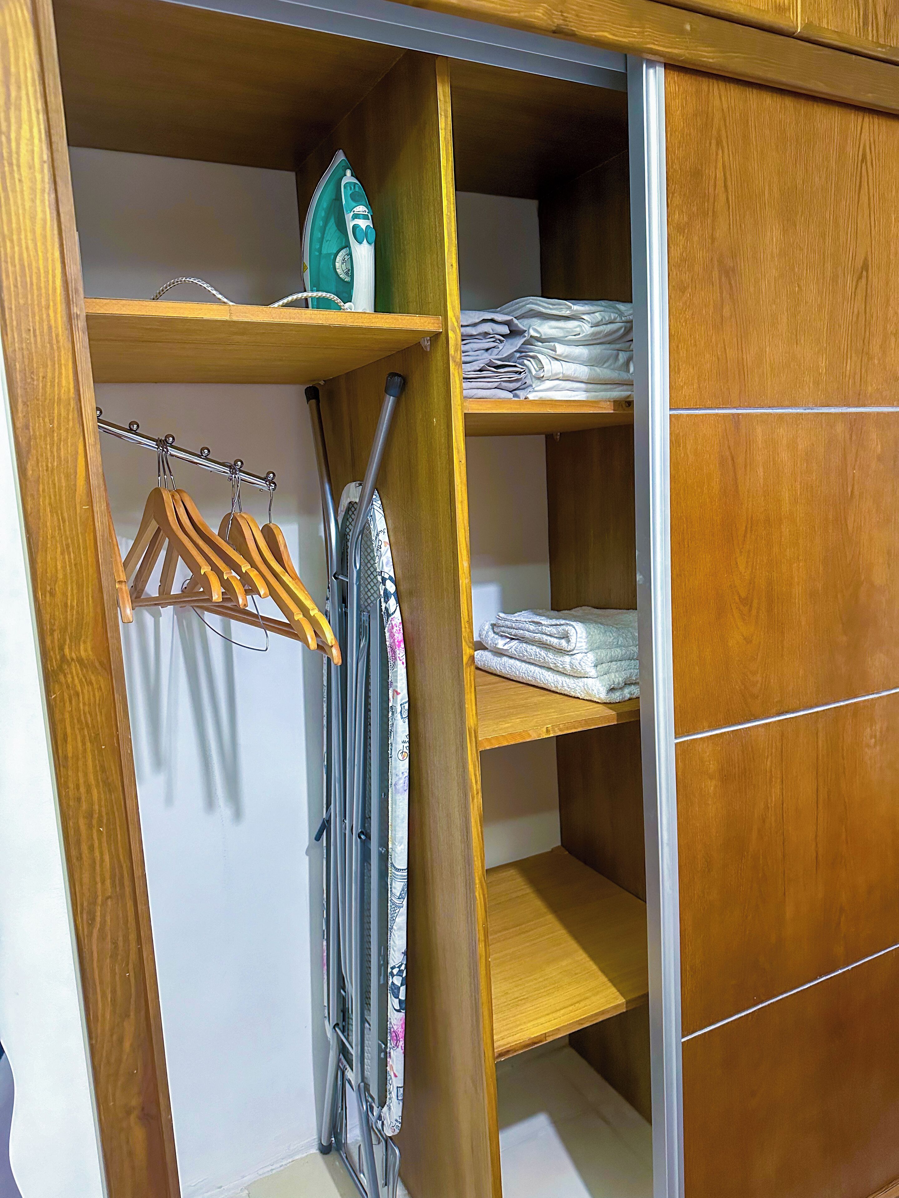 A wooden wardrobe with a mirror, a hanging rack with clothes, and a folded towel on the top shelf.
