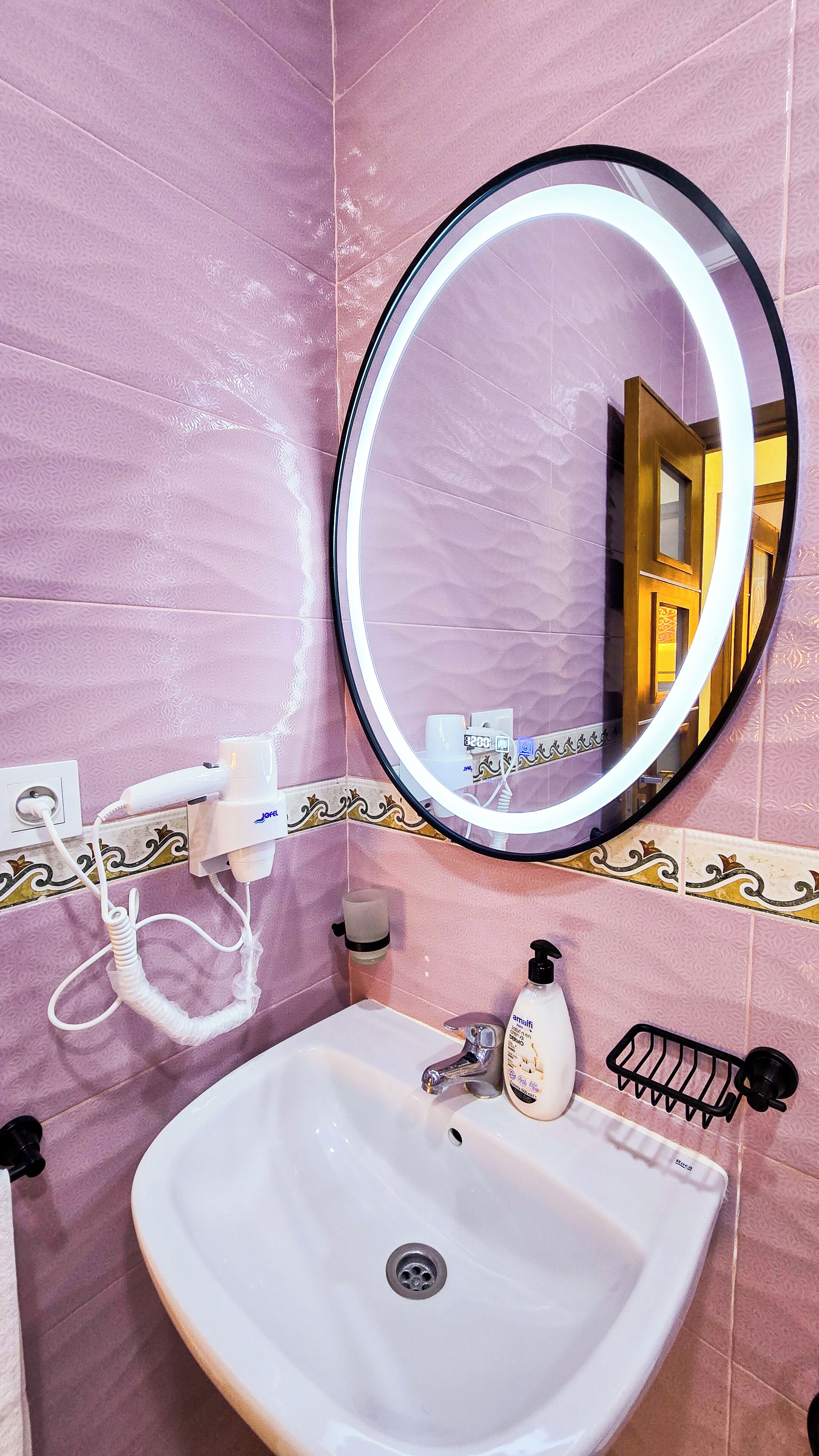 A bathroom with a white sink, a round mirror with a light, and pink patterned tiles.