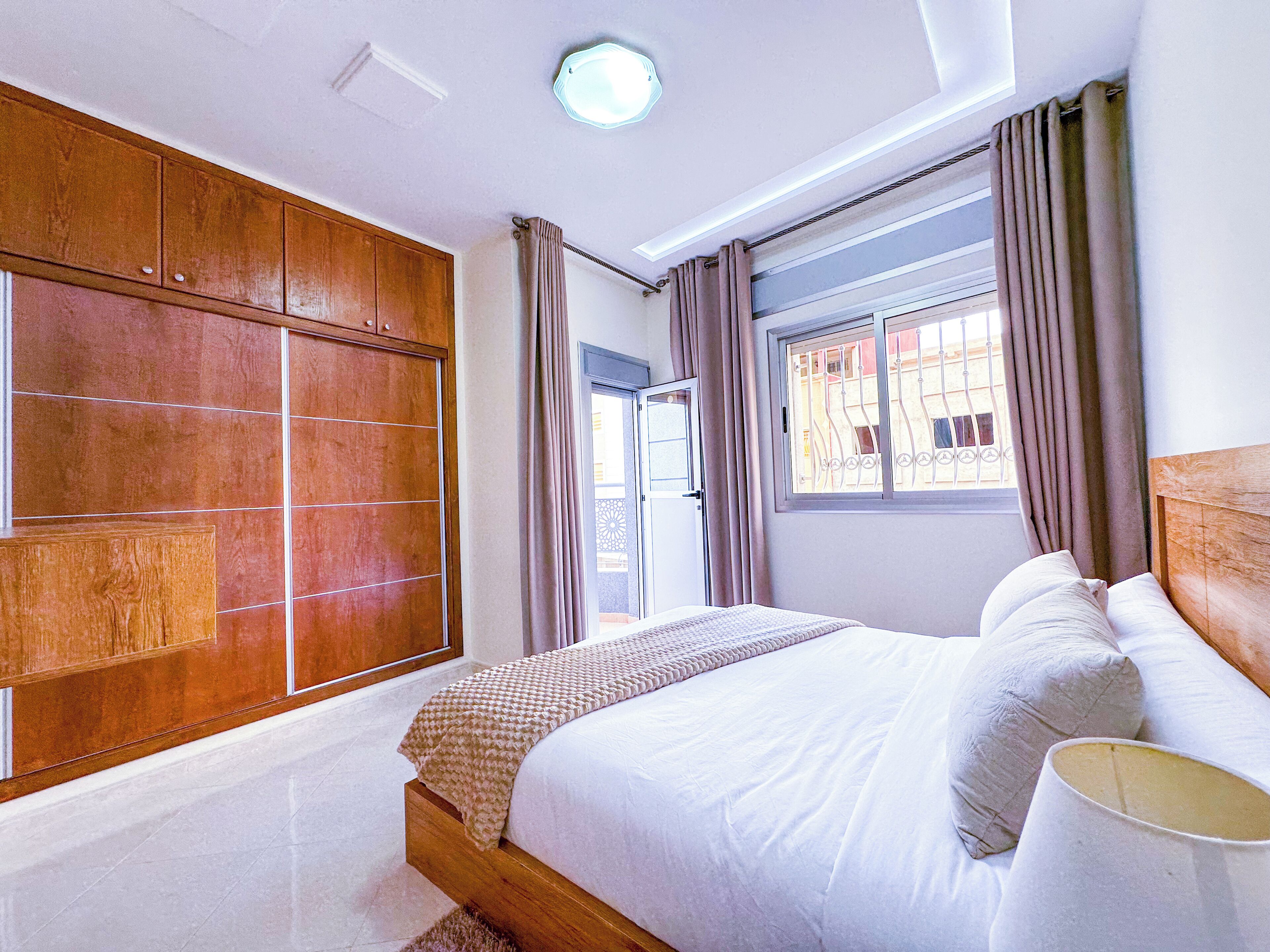 Un dormitorio con un amplio armario de madera, una cama con ropa de cama blanca, una ventana con cortinas y una lámpara en el techo.
