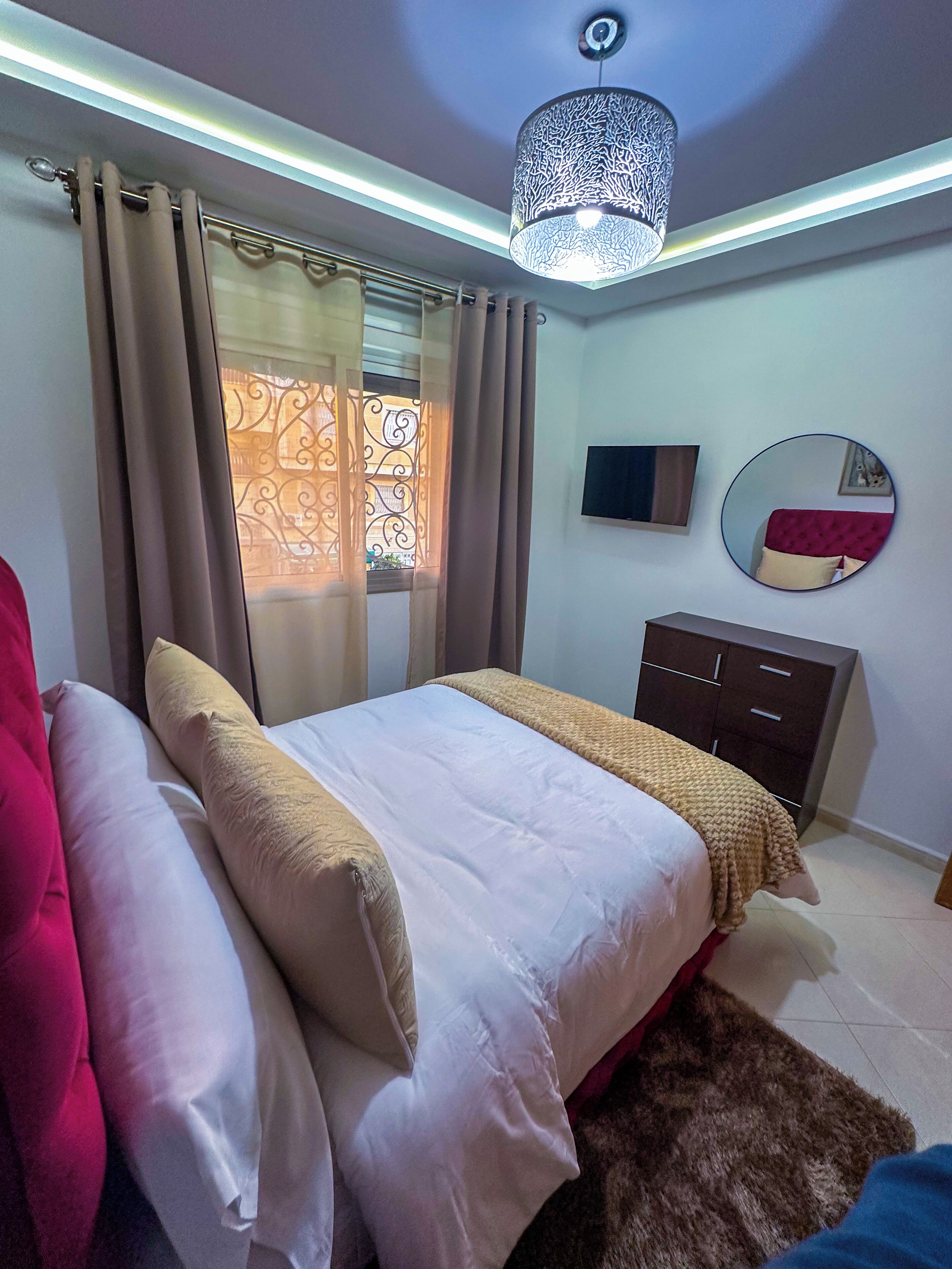 Un dormitorio con cama, cortinas, espejo, cómoda y lámpara en el techo.