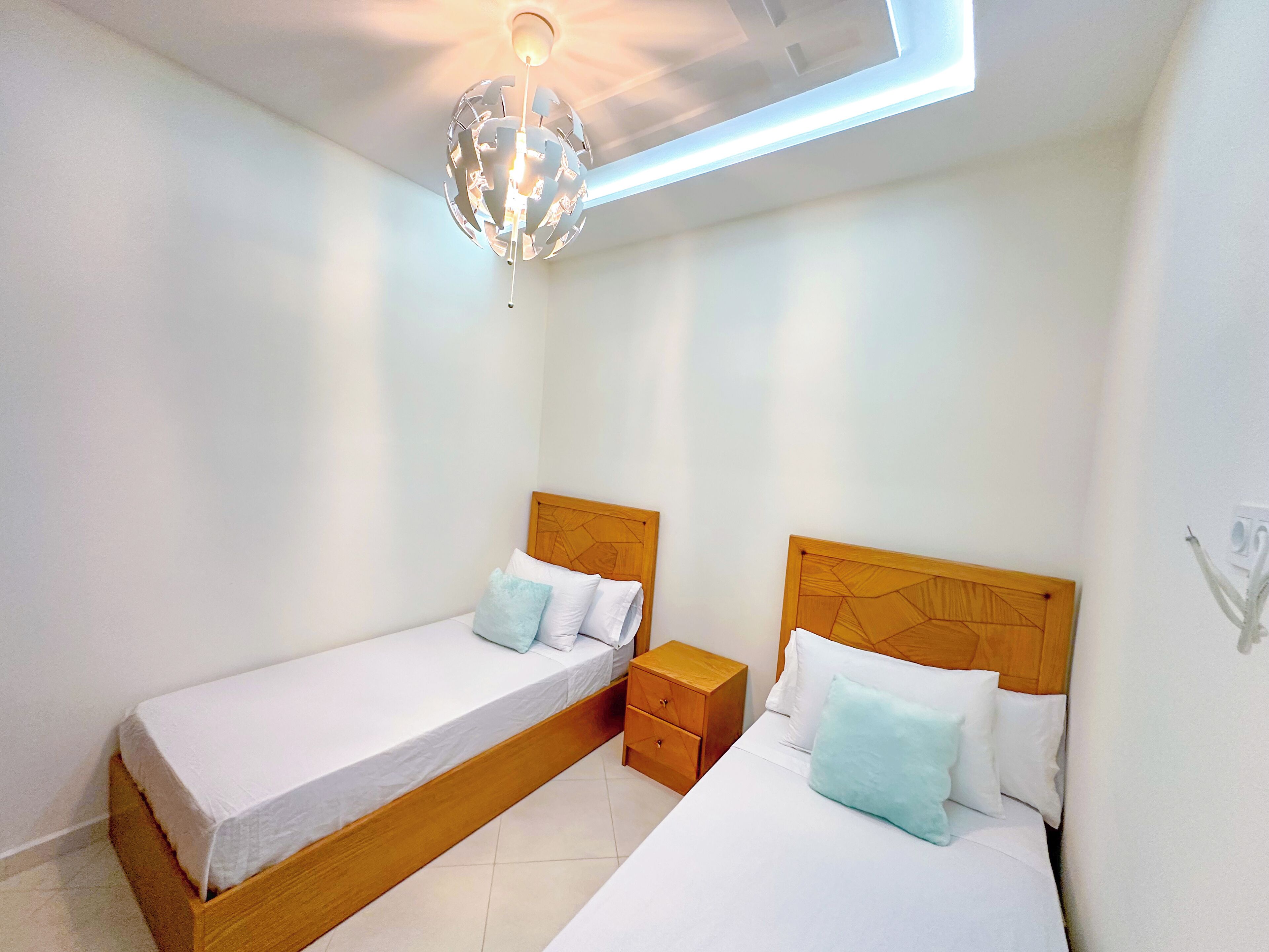 A bedroom with two single beds, wooden headboards, a nightstand, and a modern chandelier.