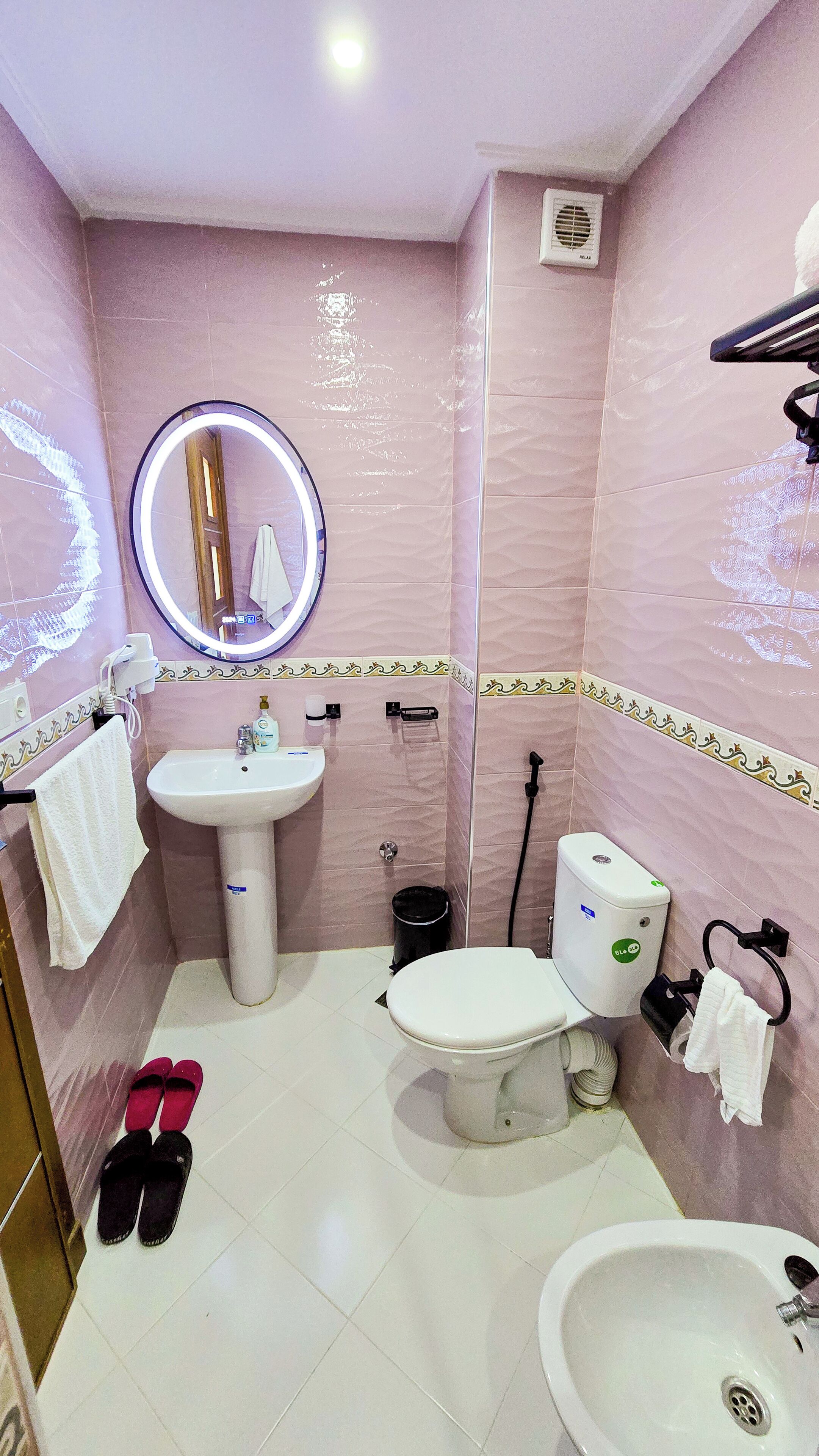 Une salle de bain équipée de toilettes, d’un bidet, d’un lavabo et d’un miroir avec éclairage. Des serviettes et des chaussons sont posés sur le sol.
