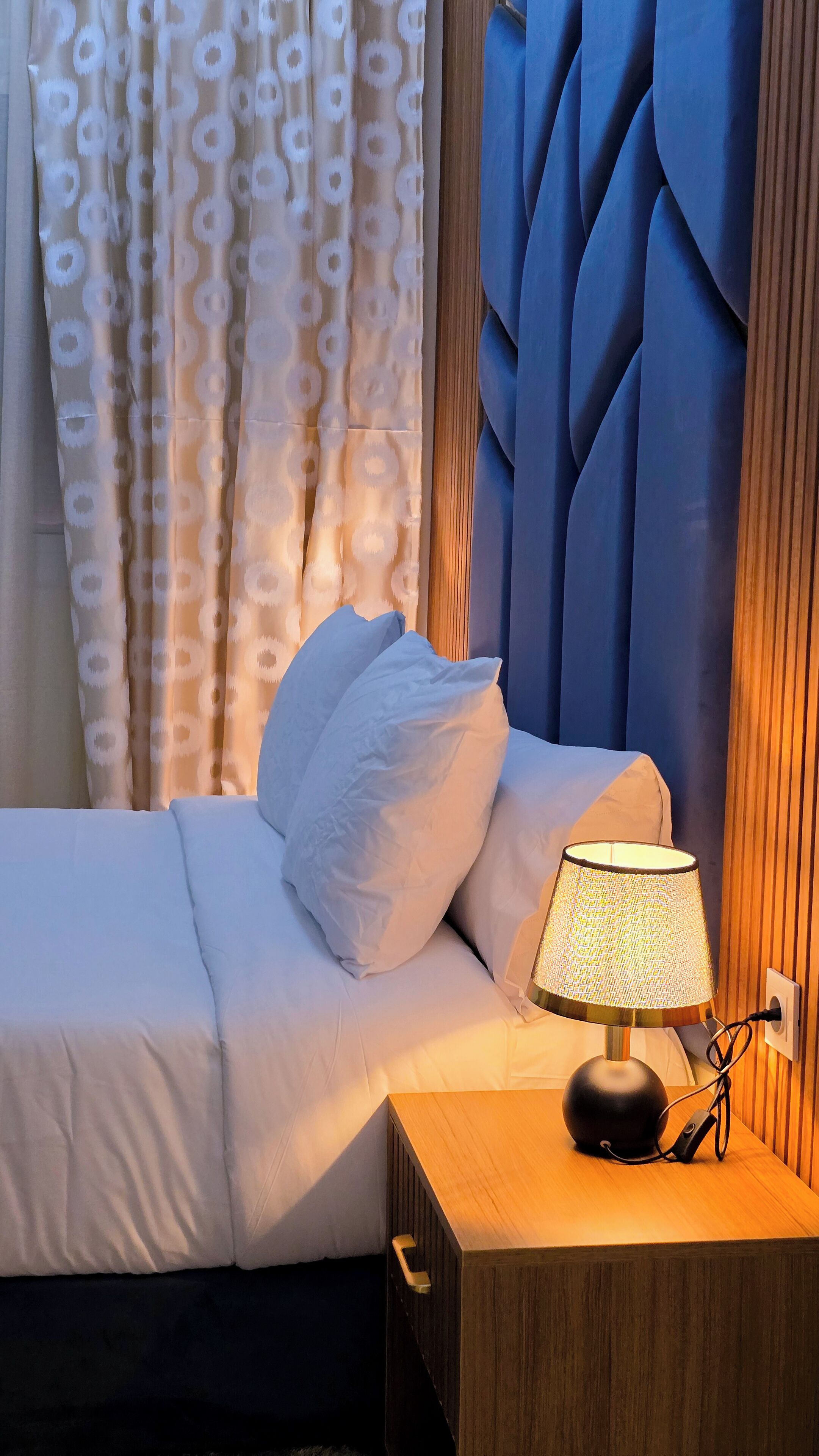 Ein ordentlich bezogenes Bett mit weißer Bettwäsche, einer Nachttischlampe und einem Holfnachtkästchen.