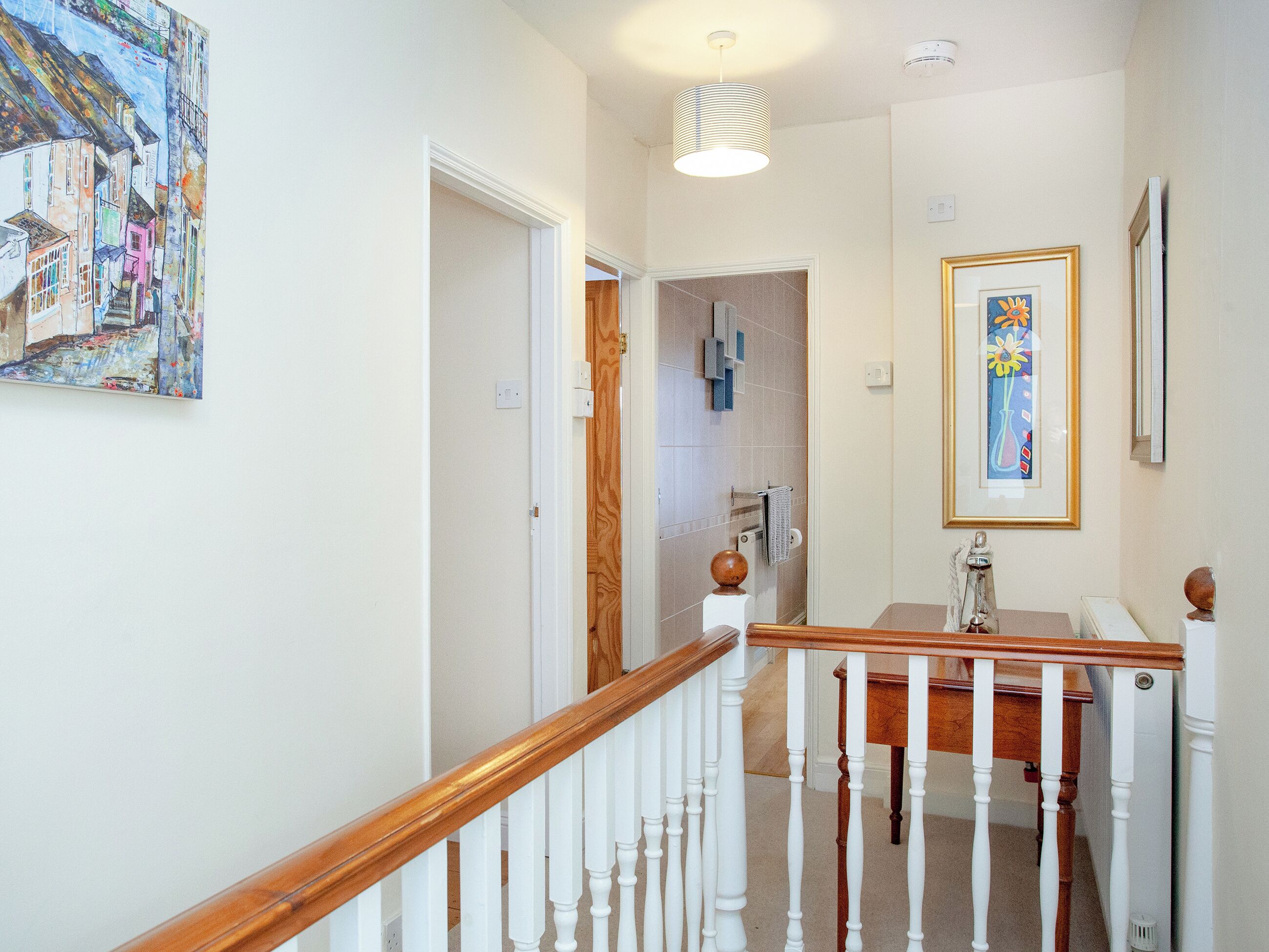 A hallway with a wooden staircase, a painting on the wall, and a framed picture above a mirror.