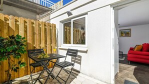 Eine kleine Terrasse mit Tisch und Stühlen, ein Holzzaun und Blick auf ein rotes Sofa in einem Zimmer.