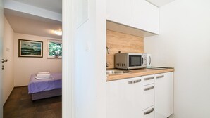 Eine kleine Küche mit weißen Schränken, einer Mikrowelle und einem Spülbecken. Im angrenzenden Raum ist ein Bett mit einer violetten Decke zu sehen.