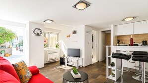 Ein modernes Wohnzimmer mit einem roten Sofa, einem Fernseher und einem kleinen Tisch mit Pflanzen. Es gibt eine Küchenzeile mit weißen Schränken und einem Barhocker.
