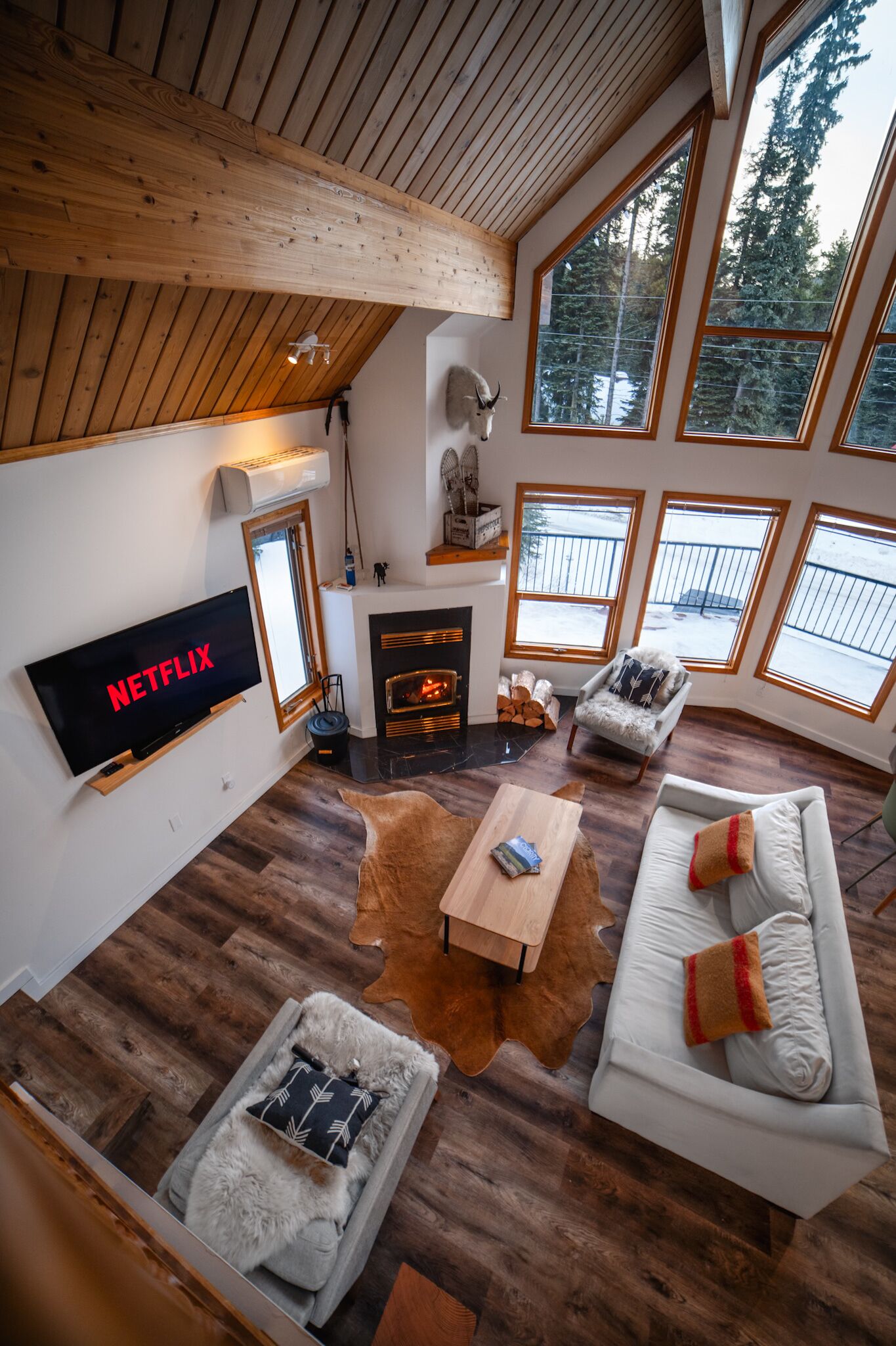 A cozy living room with a fireplace, a TV mounted on the wall, and large windows offering a view of the outdoors.