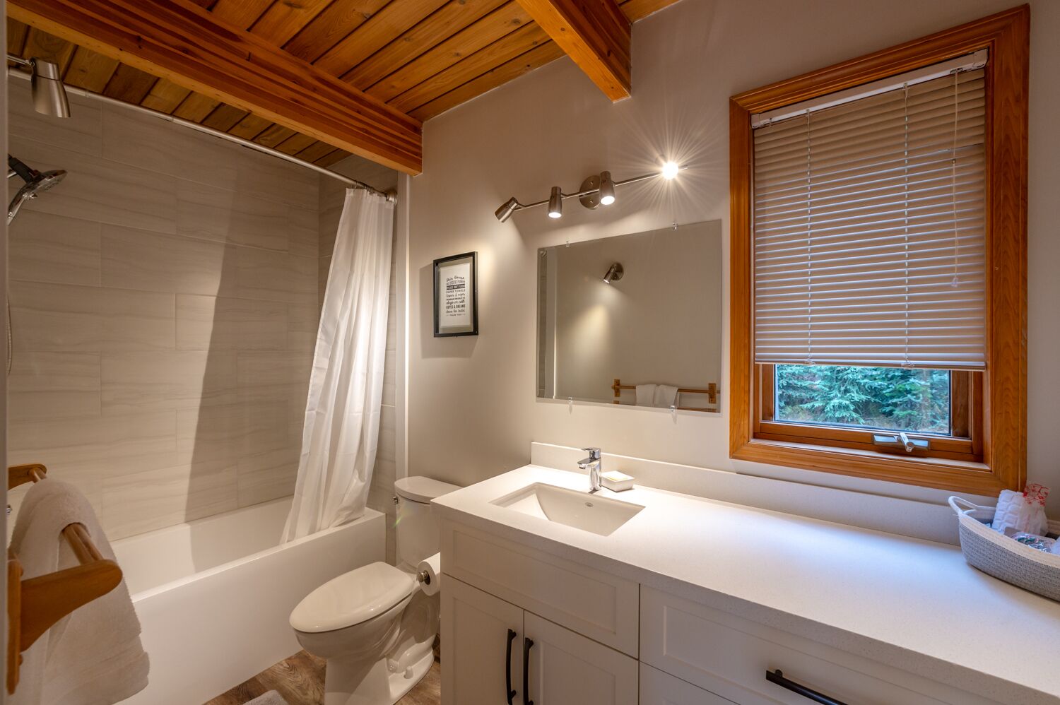 Une salle de bain moderne avec une baignoire, des toilettes et un lavabo. Il y a une fenêtre avec des stores, un rideau de douche et un tableau encadré au mur.