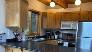 Cocina moderna con armarios de madera, encimera negra, refrigerador de acero inoxidable, microondas y cafetera. Hay tres taburetes de madera y una ventana con vistas a árboles.