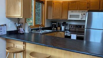 A modern kitchen with wooden cabinets, a black countertop, a stainless steel refrigerator, a microwave, and a coffee maker. There are three wooden stools and a window with a view of trees.