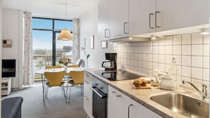 Une cuisine moderne avec des armoires blanches, un évier en acier inoxydable et une machine à café. On y trouve un coin repas avec des chaises jaunes et une table, ainsi qu’une fenêtre donnant sur des immeubles.
