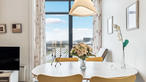 Un espace repas moderne avec une table ronde, des chaises jaunes, un vase rempli de fleurs et une vue sur la ville grâce à de grandes fenêtres.