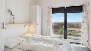 Une chambre à coucher avec un lit, une étagère ornée d’une plante, une fenêtre avec des rideaux et une vue sur un paysage verdoyant.
