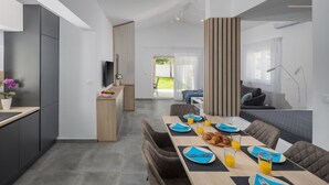 Ein moderner Essbereich mit einem langen Frühstückstisch, eine Küchenzeile mit dunklen Schränken und ein Wohnbereich mit Sofa und Fernseher.