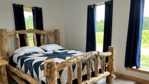 Un lit en bois rustique, recouvert d’une courtepointe, avec deux fenêtres ornées de rideaux et une vue sur la verdure.
