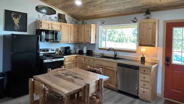 Une cuisine avec des meubles de rangement en bois, un réfrigérateur noir, une table à manger en bois et une fenêtre donnant sur un paysage verdoyant.
