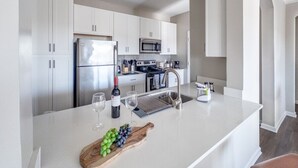 A modern kitchen with white cabinetry, stainless steel appliances, and a central island with a wine bottle and grapes. - Birkdale Pool-View, Elevator,Shop-Eat-Work-Play (Huntersville)