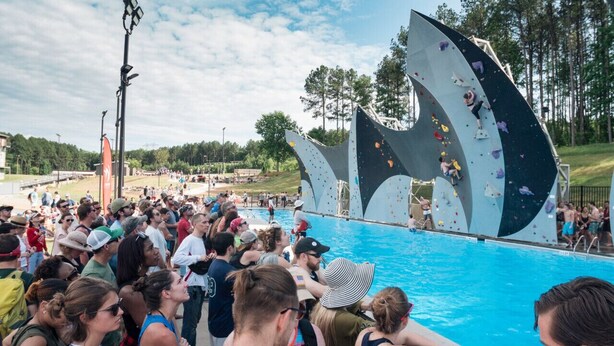 A rock climbing wall with multiple climbers at various levels, surrounded by a crowd of spectators by a pool. - Birkdale Pool-View, Elevator,Shop-Eat-Work-Play (Huntersville)