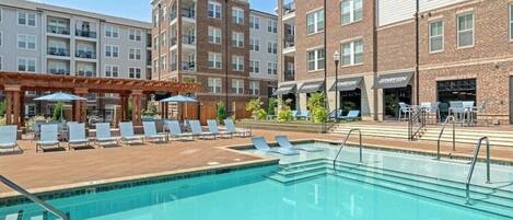 Une piscine entourée de chaises longues et de parasols, elle-même bordée d’immeubles à plusieurs étages aux façades en briques et dotés de balcons.