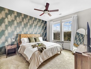 Een slaapkamer met een bed, nachtkastje, bureau, plafondventilator en raam met uitzicht.