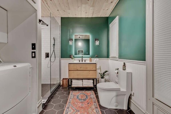 A modern bathroom with a green wall, a wooden vanity, a white toilet, and a shower area.
