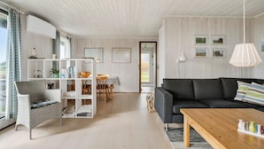 Un salon spacieux doté d’un plafond en bois, d’un canapé gris foncé, d’une table basse en bois et d’une étagère blanche où sont disposés des livres et des objets décoratifs.