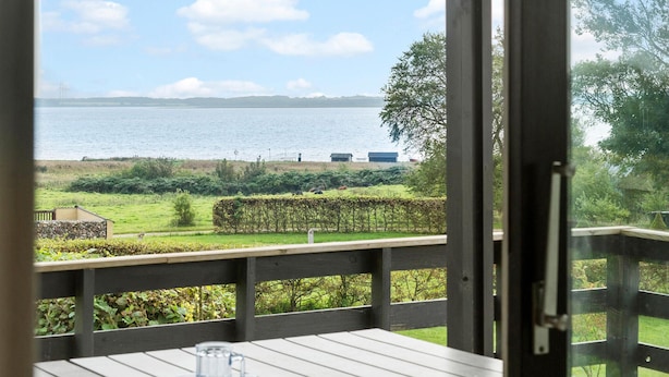 Une terrasse en bois avec une table et des chaises, offrant une vue sur une étendue d’eau, une zone herbeuse et des éoliennes situées au loin.