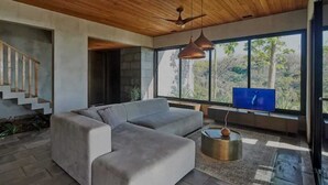 Een moderne woonkamer met een hoekbank, een televisie en een groot raam met uitzicht op bomen.