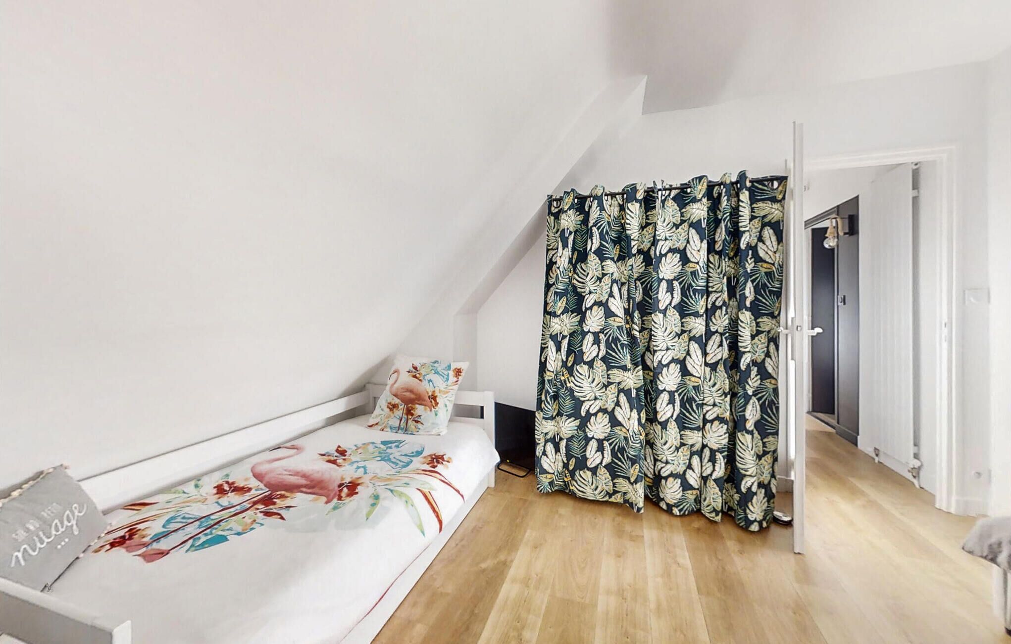 A bedroom with a bed, a nightstand, a floral-patterned pillow, and a tropical leaf-patterned curtain.