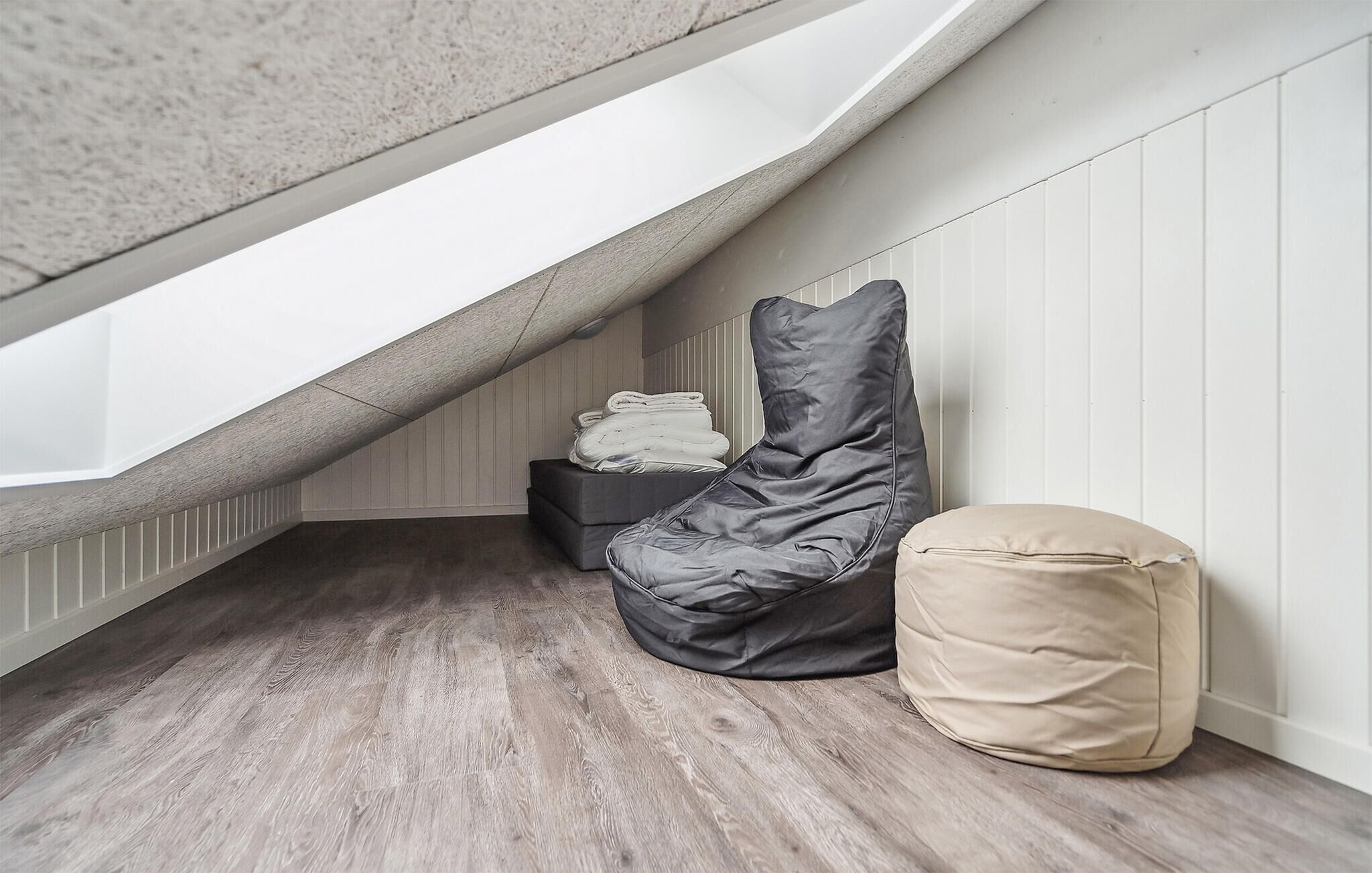A loft bedroom with a sloped ceiling, a grey bean bag chair, a beige storage bag, and a folded blanket on a wooden floor.