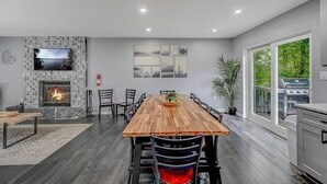 A modern dining area with a wooden table, chairs, a fireplace, and a large window overlooking greenery. - Pocono Paradise: Spacious Fun for the Whole Family (Long Pond)