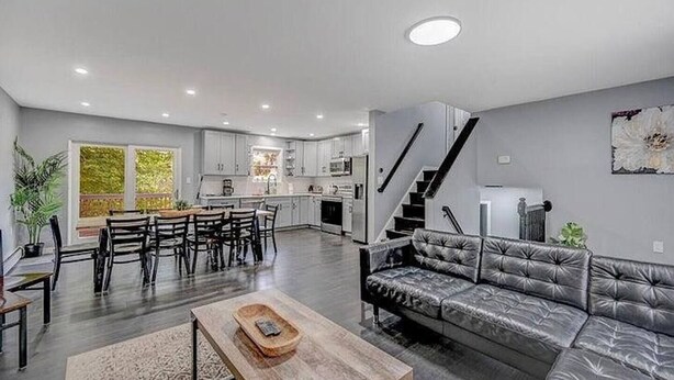A modern kitchen with white cabinets, a dining area with a wooden table and chairs, a gray leather sofa, and a staircase leading to another level. - Pocono Paradise: Spacious Fun for the Whole Family (Long Pond)