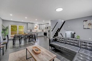 A modern kitchen with white cabinets, a dining area with a wooden table and chairs, a gray leather sofa, and a staircase leading to another level. - Pocono Paradise: Spacious Fun for the Whole Family (Long Pond)