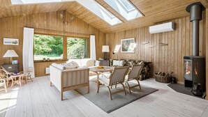 Interior acogedor de una cabaña con paredes de madera, un sofá, sillas, una mesa, una chimenea y ventanas amplias que ofrecen vistas al exterior.