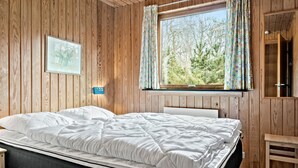 Una habitación con paneles de madera, una cama, un ventanal con cortinas, una mesita de noche y un pequeño banco.