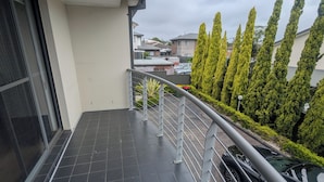 Un balcón con suelo de baldosas, baranda de metal y vista a edificios residenciales y árboles.